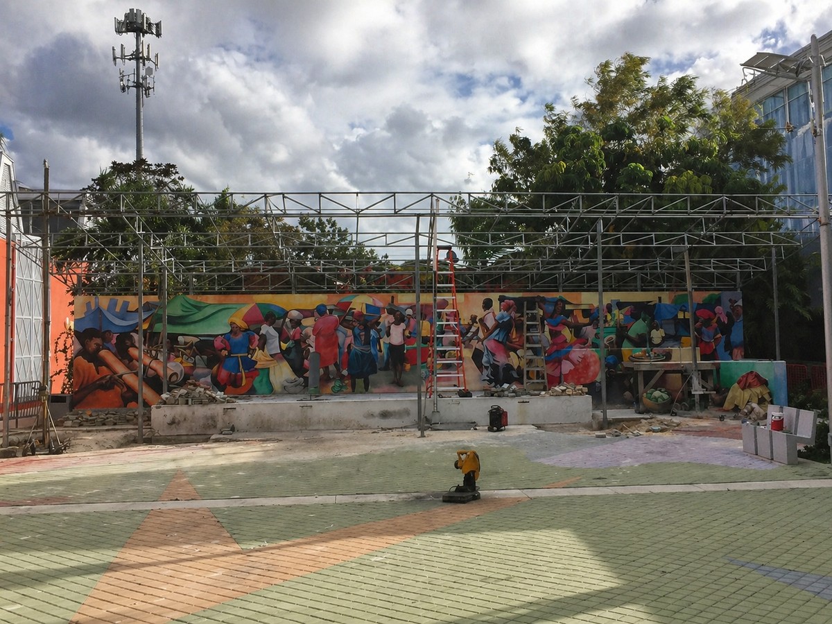 Haitian Cultural Center plaza with bare metal canopy frame mid-installation, scaffolding visible, colorful Haitian mural and modern glass building behind.