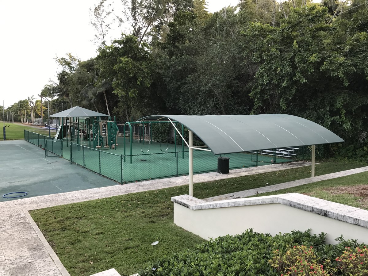 Wide view of arched green-canopy spectator shelter beside a tennis court enclosed in dark mesh fencing with mature tree canopy.