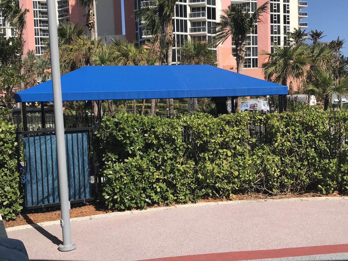 Royal-blue hipped pavilion canopy at a coastal high-rise condo amenity area, sea-grape hedge in the foreground and pink towers behind.