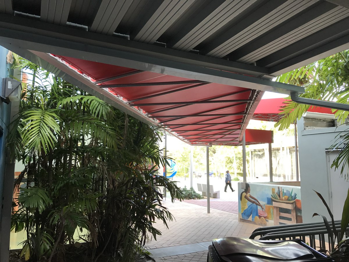 Underside of a red truss-frame canopy with palm fronds in foreground at the Wynwood-style cultural center, person walking in the distant courtyard.