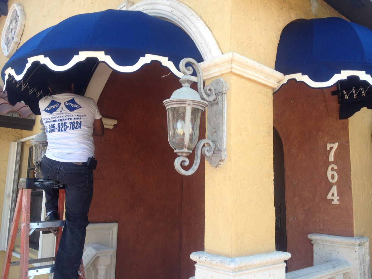 Crew member in Awning Brokers branded white tee installs a navy scalloped dome awning over the arched entry of a Mediterranean home.