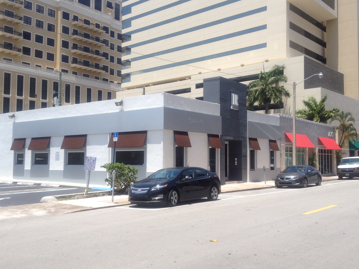 Long Brickell-area single-story commercial building with row of small burgundy projecting window awnings tucked beneath modern high-rise towers.