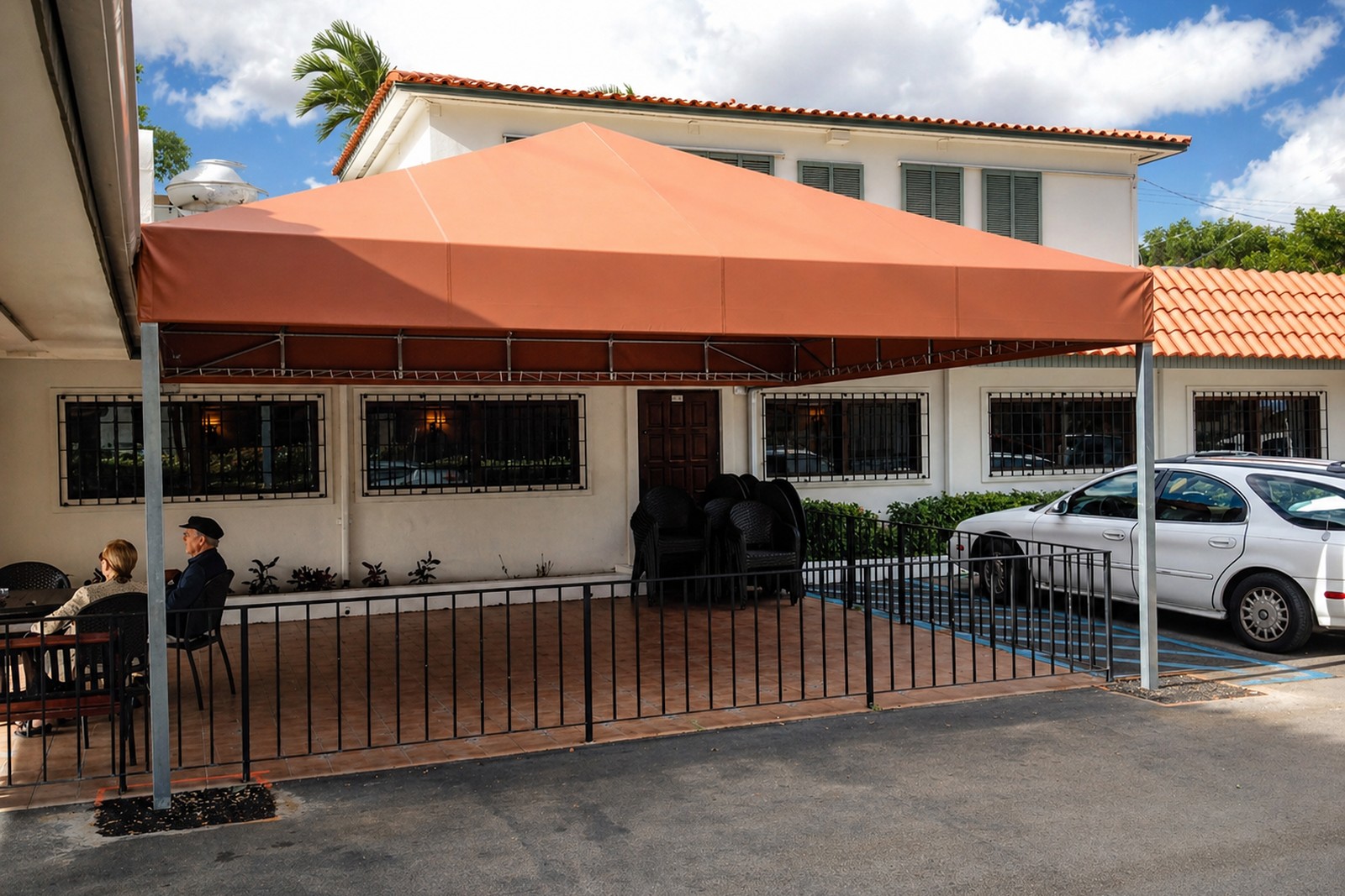 Tall terra-cotta hipped patio canopy on slim aluminum posts shades a small outdoor restaurant patio with iron rail and tile-roof building beyond.
