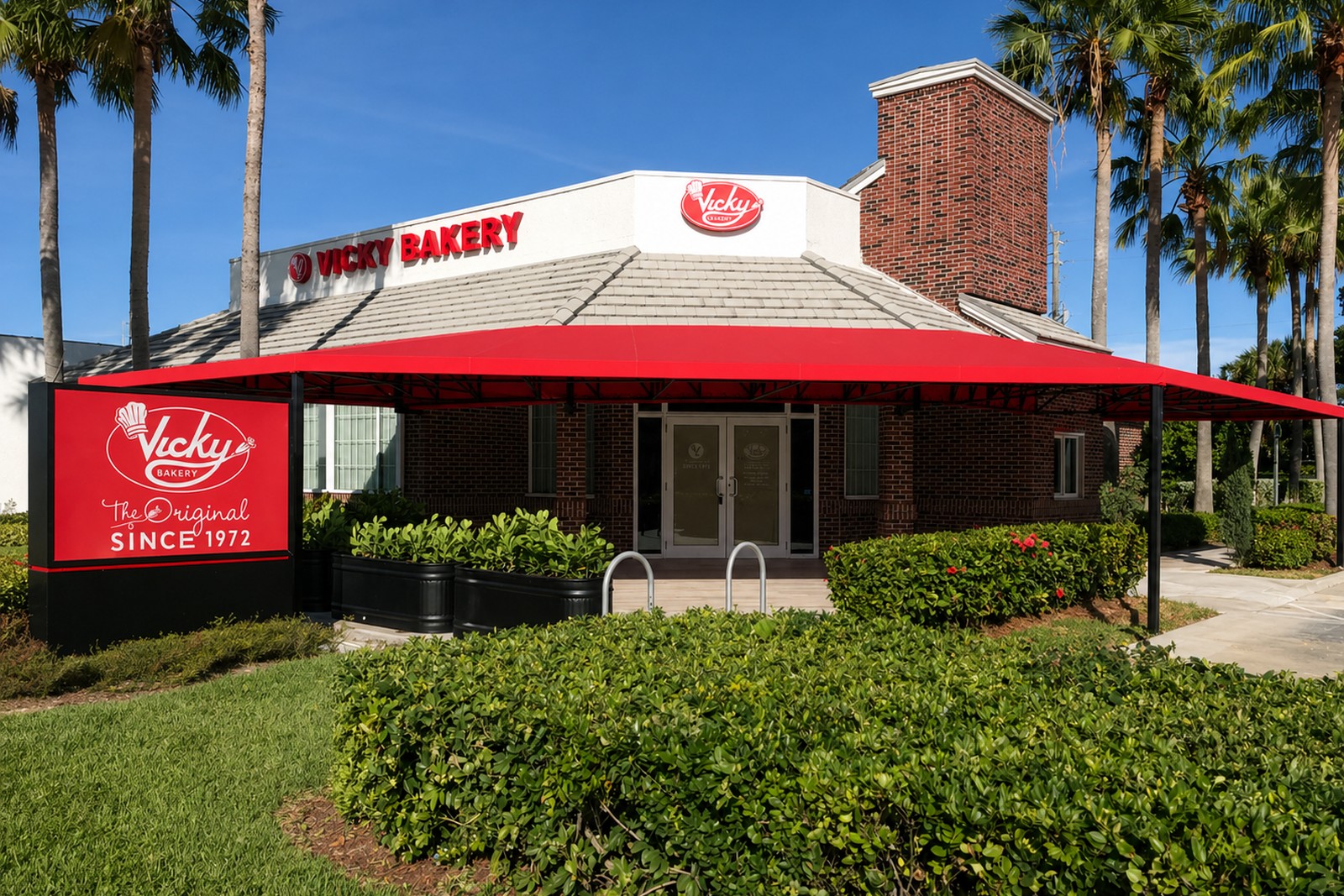 Vickys Bakery storefront wraps a hipped red canopy with white piping over the brick facade, palms framing the entrance against a clear sky.