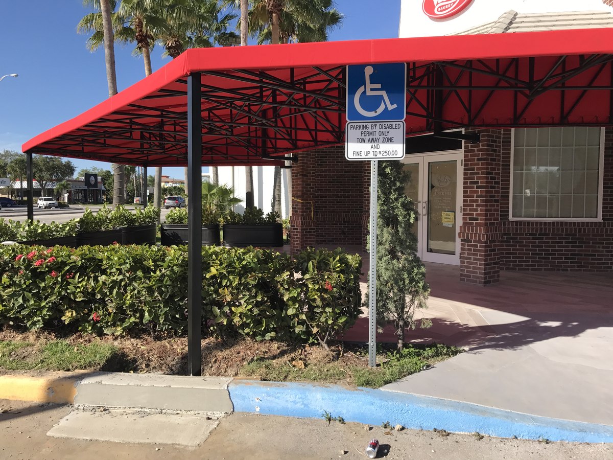 Side wing of Vickys Bakery with a long red awning over the sidewalk, ADA accessible parking sign and brick column accent.
