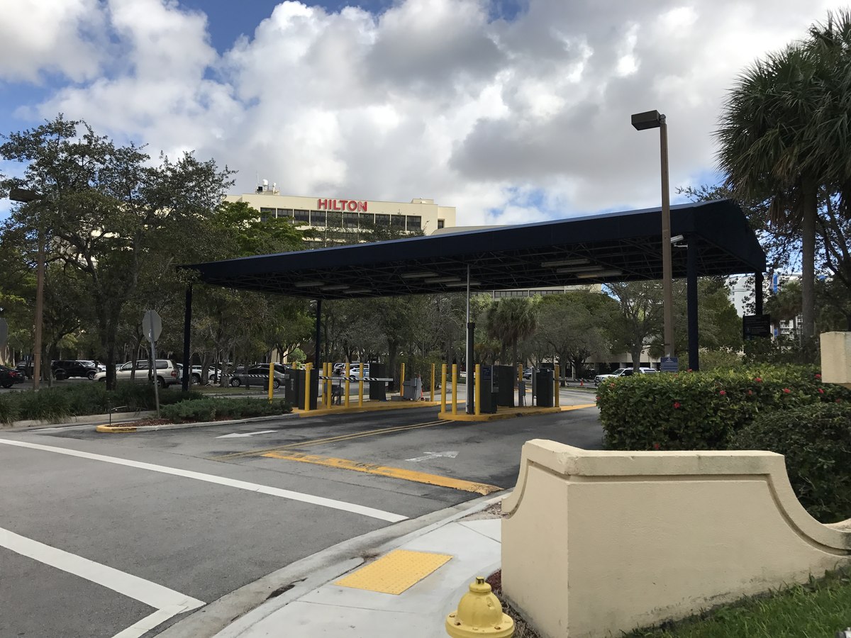Wide approach showing the full navy entry canopy of the Hilton with stucco gate post, palms and the Hilton tower beyond.