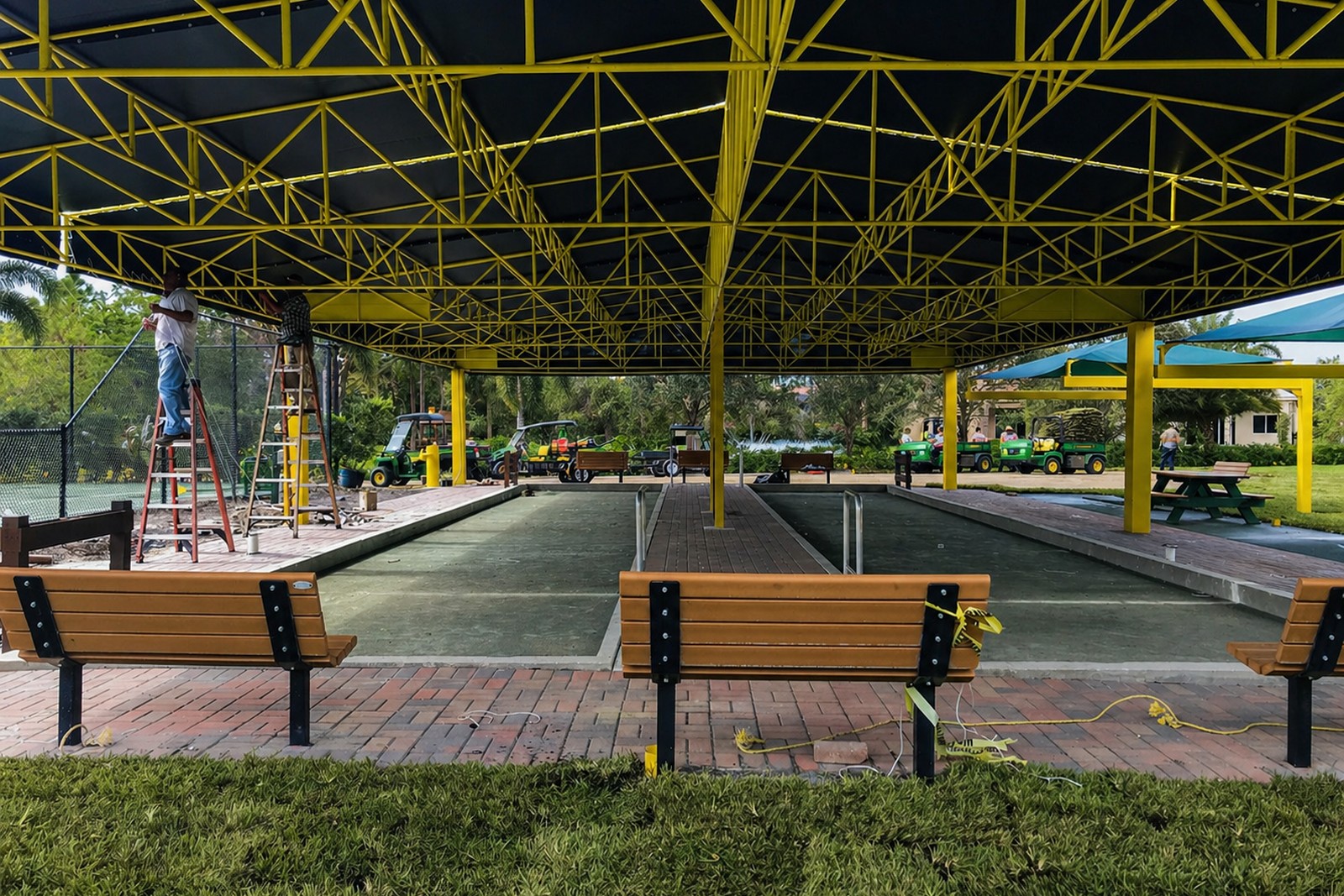 Underside of large shade canopy with steel framing, yellow posts, and benches lining bocce-court alleys at a community park.