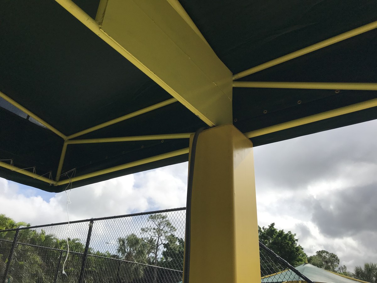 Architectural detail of the yellow truss column meeting the green canopy underside, chain-link fence and trees beyond.