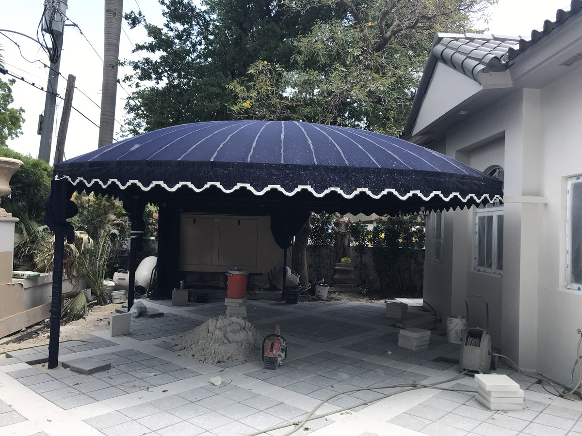 Navy dome awning with white scallop trim over a freshly-paved courtyard at a Mediterranean home, masonry tools and sand pile in foreground.