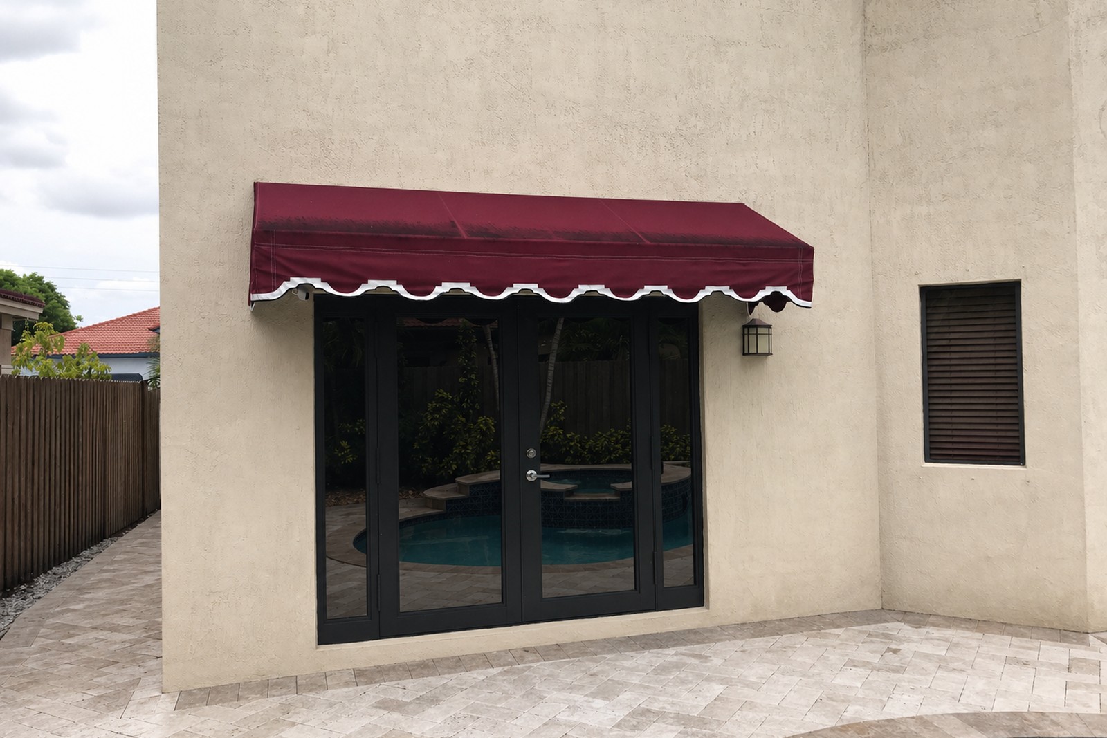 Burgundy awning with white scalloped piped trim shelters a black French-door entry to the pool patio of a tan stucco home.