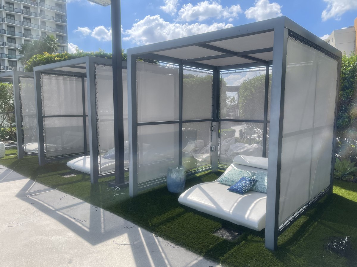 Modern aluminum-framed poolside cabanas with translucent panels and white cushioned daybeds, on artificial-turf deck.