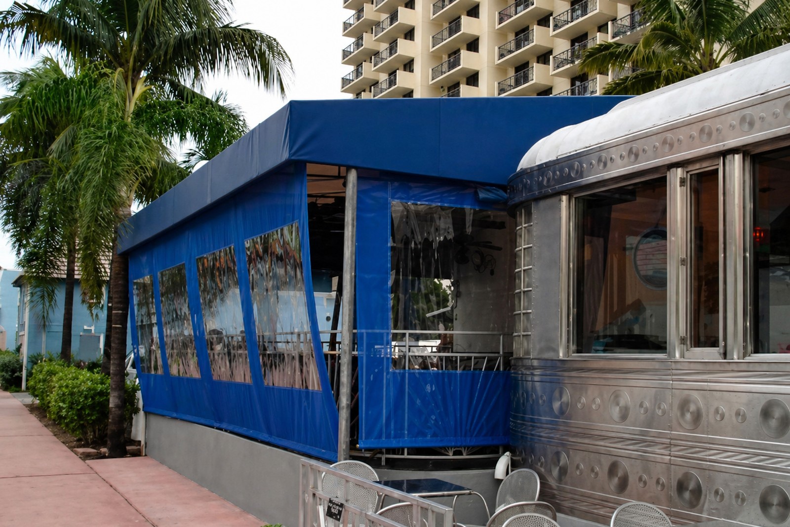 Bold blue valance canopy with clear roll-down storm panels wraps a stainless-clad diner under a Miami Beach high-rise and palms.
