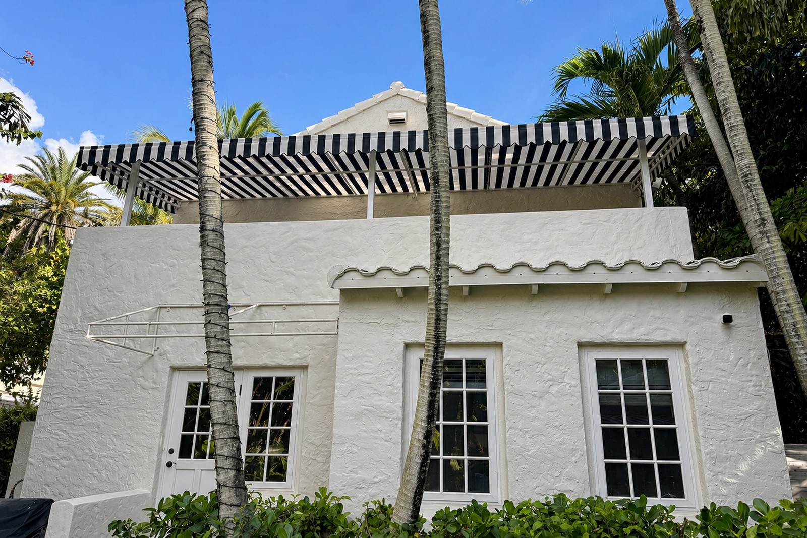 Black-and-white striped retractable awning shades a second-floor terrace at a white Mediterranean villa, palms framing the view.
