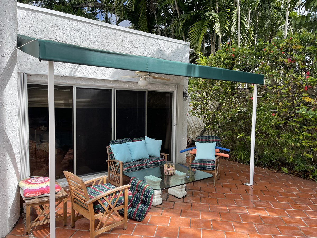 Forest-green retractable lateral-arm awning shades a terra-cotta tile patio with rattan loveseat and bistro table, palm canopy behind.