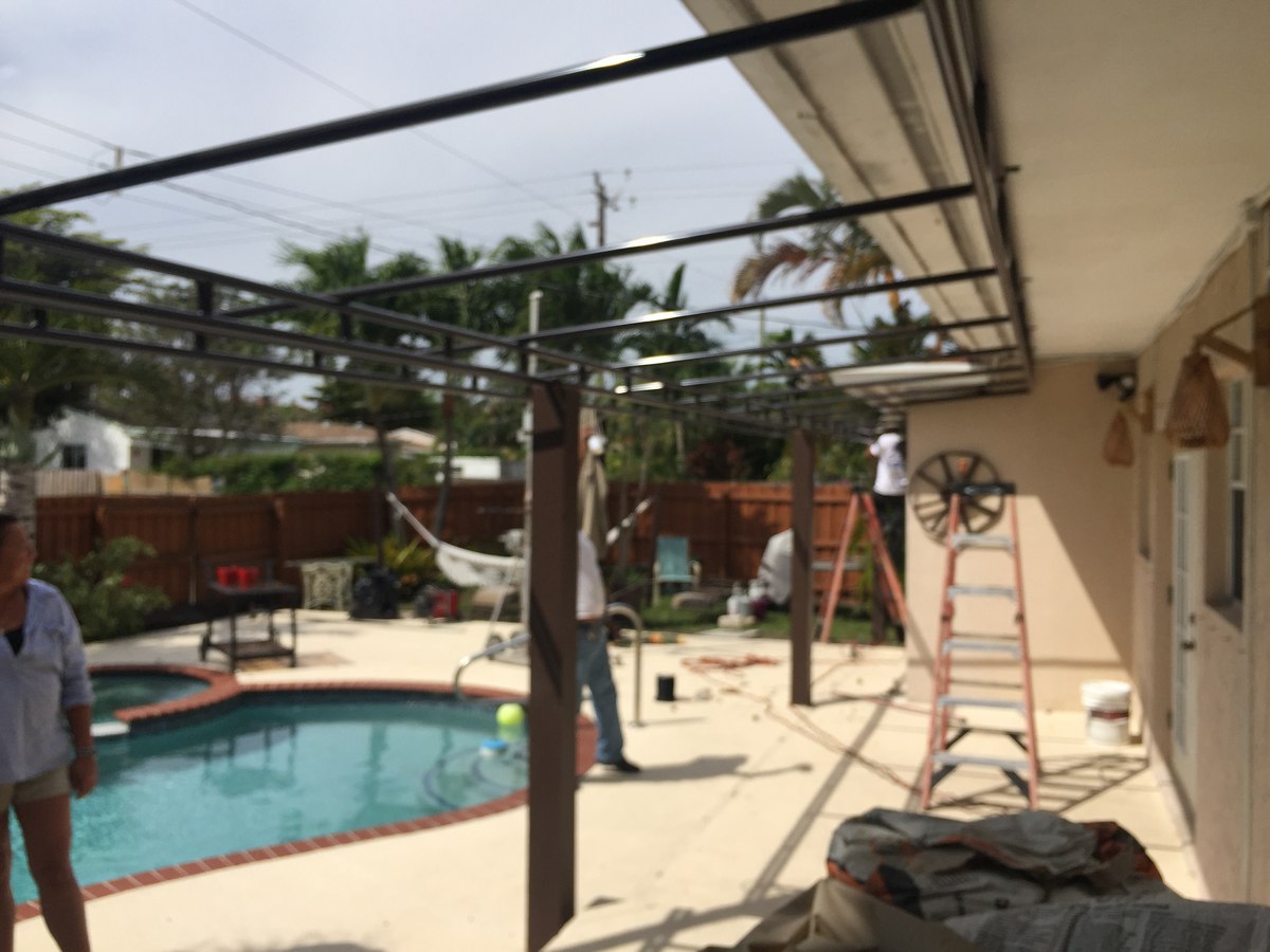 Steel pergola/awning frame mid-installation over a residential pool patio, two workers and a stepladder visible at the wall side.
