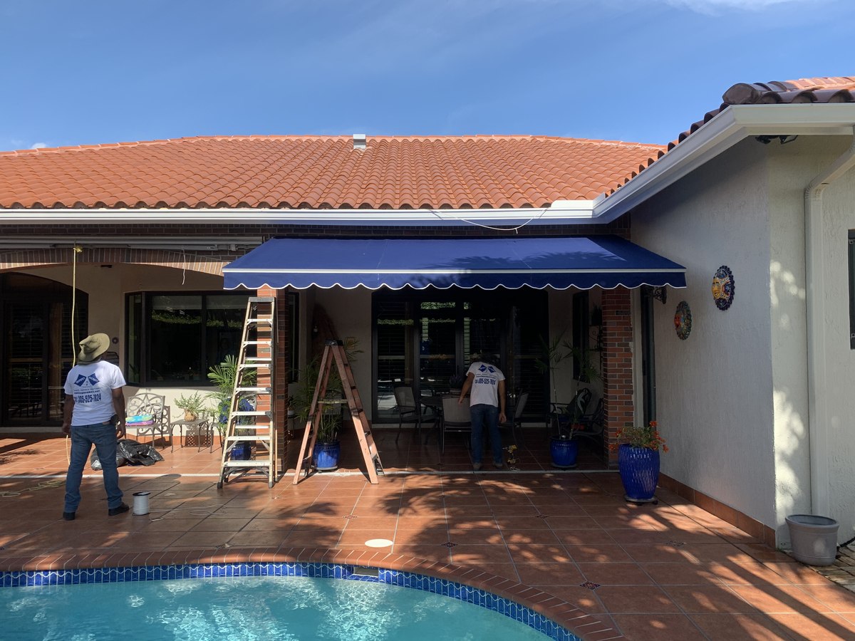 Awning Brokers crew in branded blue shirts (305-525-7824) installing a navy retractable awning over a tile-roof Mediterranean patio with pool.