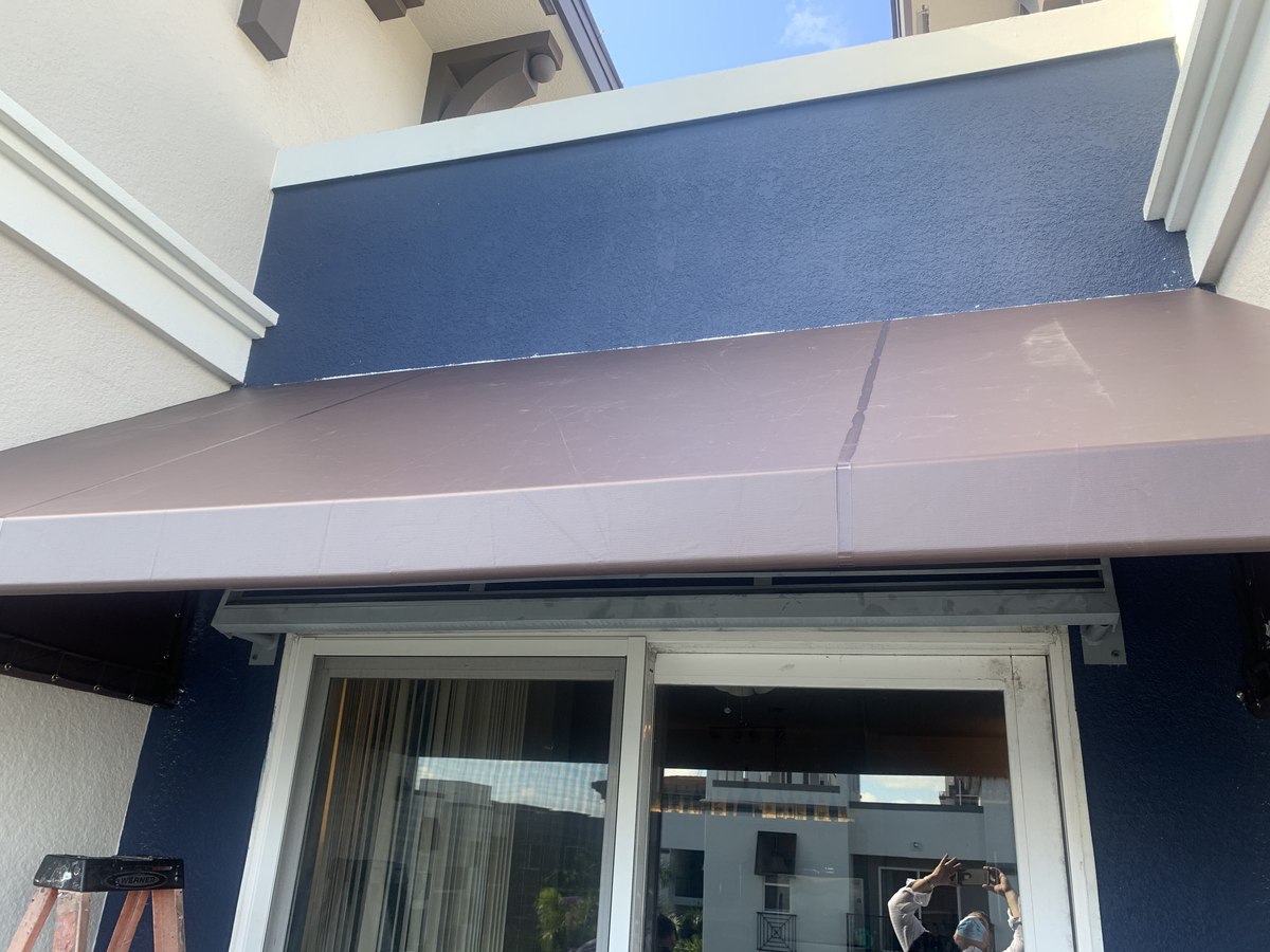 Newly installed cocoa-canvas fixed awning over a sliding glass door on a navy-painted commercial facade with a step ladder in frame.