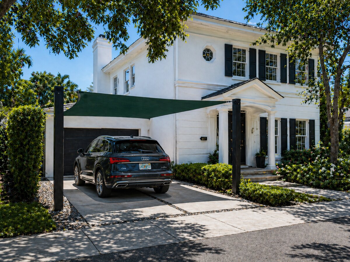 A forest-green shade sail covering one vehicle next to a classic colonial home with black shutters