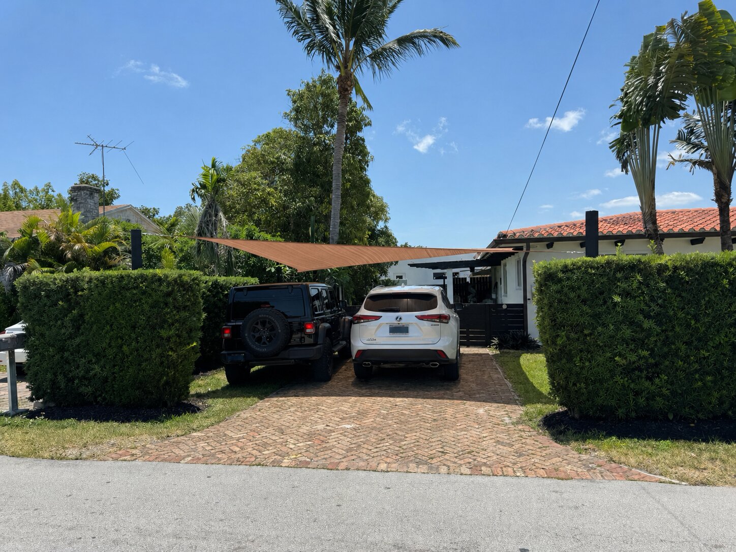 The same shade sail covering two vehicles &mdash; a Jeep Wrangler and a Toyota Highlander &mdash; in a brick-paver South Florida driveway