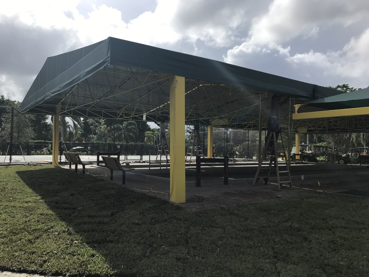 Massive yellow steel-truss park canopy with green fabric panels under installation over a bocce court area with crew on ladders.