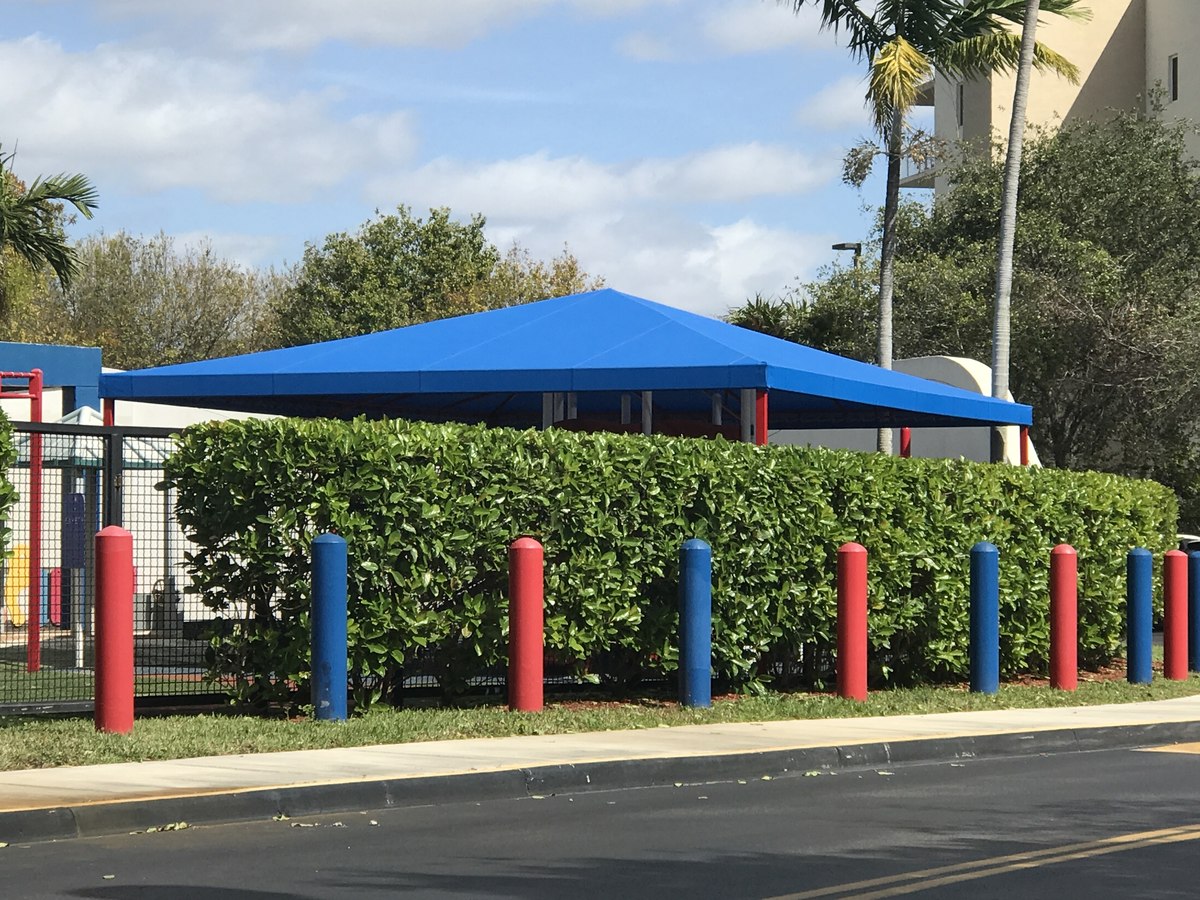 Bright royal-blue hip-roof shade canopy at a school playground, framed by red and blue bollards and a hedge along the sidewalk.