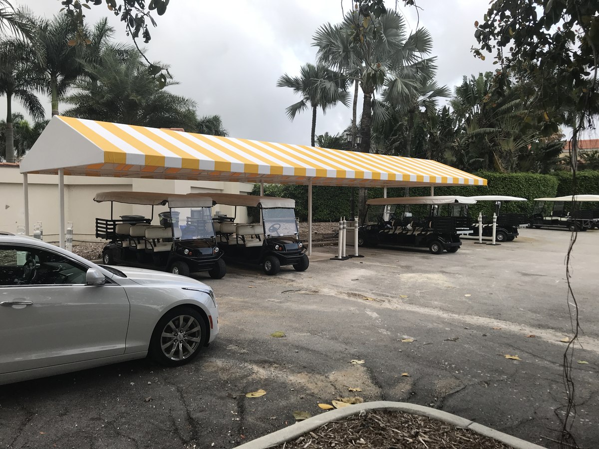 Long yellow-and-white striped golf-cart canopy on white frame at a tropical private club, palms behind and silver Cadillac in foreground.
